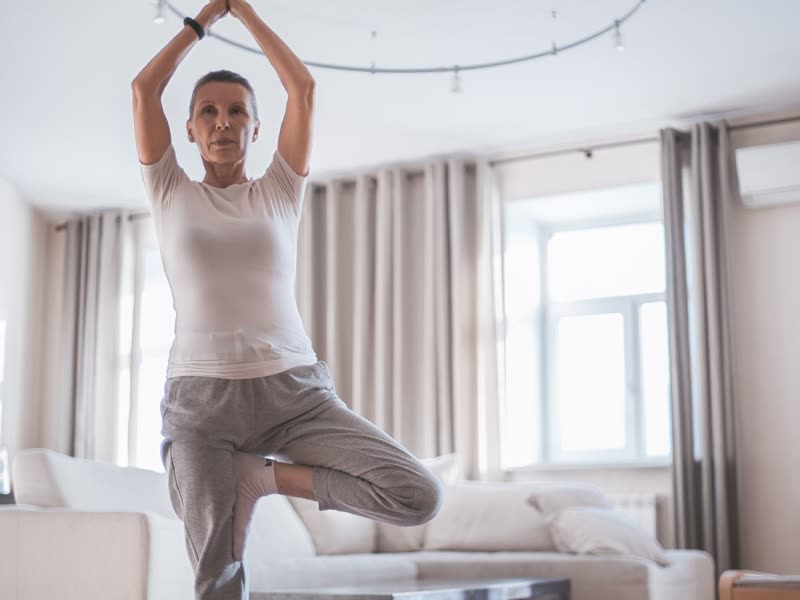 A woman balancing on one leg in her living room, arms overhead.